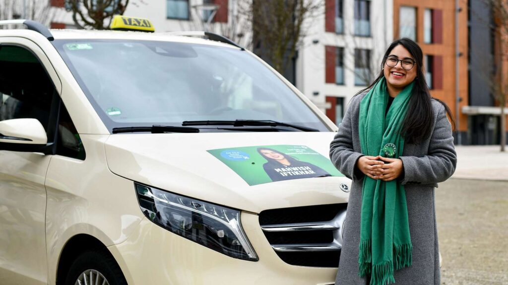 Auf dem Bild ist Mahwish Iftikhar abgebildet, sie steht lächelnd vor einem Taxi.