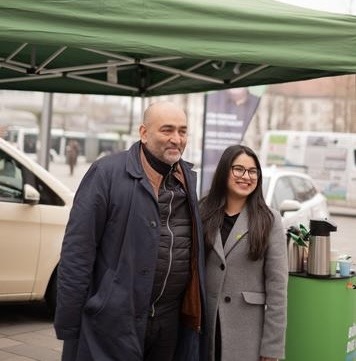 Auf dem Foto sind Omid Nouripour (links) und Mahwish Iftikhar (rechts) lächelnd Arm in Arm vor einem Taxi abgebildet