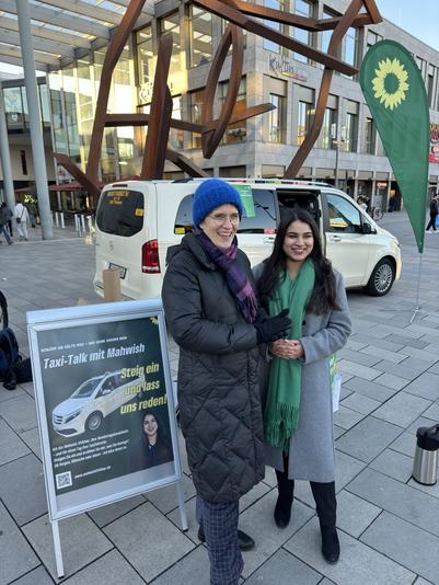 Auf dem Foto sind Anna Lührmann (links) und Mahwish Iftikhar (rechts) lächelnd Arm in Arm vor einem Taxi abgebildet. 